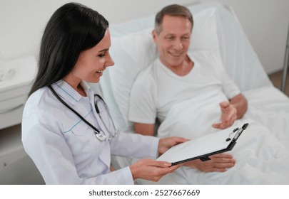 Smiling female doctor reviews notes on clipboard with a male patient lying in a hospital bed. The patient is engaged and relaxed. The bright, clean room emphasizes professional care and communication - Powered by Shutterstock