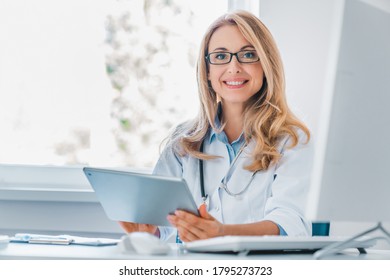 Smiling Female Doctor Sitting At Her Desk Holding Digital Tablet