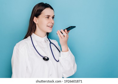 Smiling Female Doctor Wearing White Medical Uniform And Stethoscope Holding Phone Speak Activate Virtual Digital Voice Assistant On Smartphone, Consult Patient Online, Isolated On Blue Background