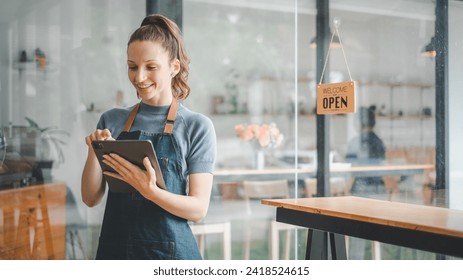 Smiling female employee in a denim apron using a digital tablet in a cozy cafe with an 'Open' sign in the background.

 - Powered by Shutterstock