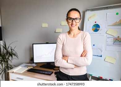 Smiling Female Employee In Glasses Looks In Camera In Office And Standing Straight In Front Of An Office Desk. A Young Successful Woman With Crossed Arms Stands Near Flipchart, Graphs On Background.