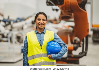 Smiling female engineer in safety gear holding a hard hat inside an industrial factory. - Powered by Shutterstock