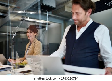 Smiling Female Entrepreneur Seen Through Glass Shield By Colleague While Working At Creative Office During COVID-19 Pandemic