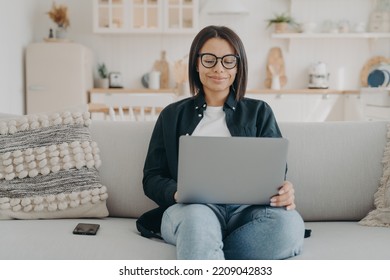 Smiling Female Freelancer Wearing Glasses Working On Laptop Sitting On Sofa At Home. Busy Young Woman Check Email, Social Networks, Shopping Online On Computer. Remote Job, E-commerce Concept.