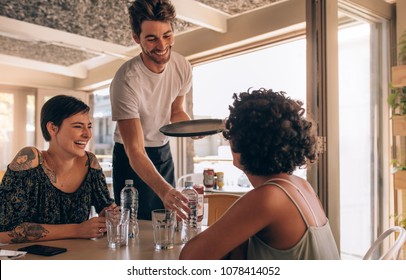 Smiling Female Friends At A Cafe With Waiter Serving Water. Two Young Women Sitting At A Restaurant.