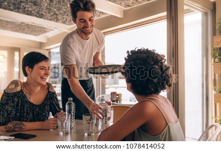 Smiling female friends at a cafe with waiter serving water. Two young women sitting at a restaurant.