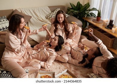 Smiling female friends enjoying pajama party at home, chatting and eating tasty pizza