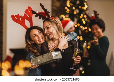 Smiling Female Friends Huging And Giving Presents To Each Other While Gathering At Home To Celebrate Christmas Or New Year.