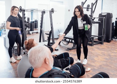 Smiling female gym worker gives freshly made cocktail to elderly client. Couple of seniors sitting in massage chairs and going to drink smoothies after electrical muscle stimulation training. - Powered by Shutterstock