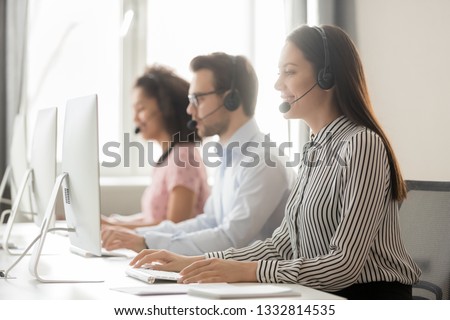 Smiling female helpline call center agent operator telemarketer in wireless headset work with colleagues in customer care support service office talking consulting client at workplace using computer