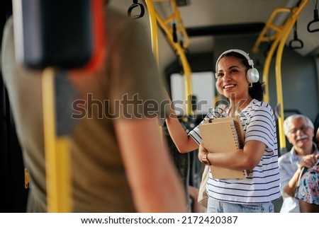 Smiling female hispanic student, holding books, listening music via bluetooth earphones and traveling by public bus