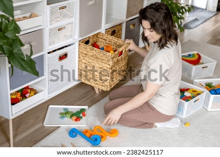 Smiling female housewife childish toys storage organizing sorting details with basket and PVC container. Woman cleaning kids room filling cabinet by multicolored playthings comfortable maintaining