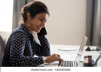 Smiling female indian employee multitask working on computer in office talking over phone, happy woman worker using laptop, laughing speaking with friend on cell or communicating with client online