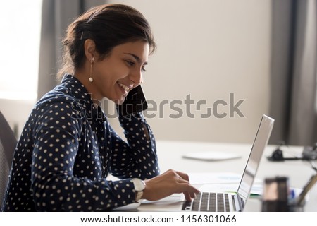 Smiling female indian employee multitask working on computer in office talking over phone, happy woman worker using laptop, laughing speaking with friend on cell or communicating with client online