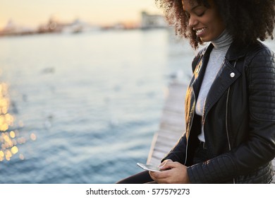 Smiling Female International Student With Curly Hair And Trendy Outfit Enjoying View Of Autumn Sunset Resting After Lesson Setting Near River Having Online Conversation With Best Friend Via Phone