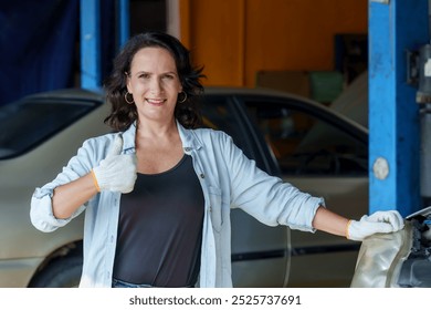 Smiling female mechanic giving thumbs-up while standing next to a car in an automotive workshop, wearing gloves and casual attire, expressing confidence in her work and successful car repair service. - Powered by Shutterstock