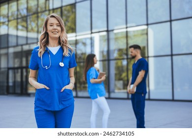 A smiling female nurse with blonde hair stands confidently outside a medical facility, her blue scrubs suggesting a professional healthcare environment while colleagues engage behind her. - Powered by Shutterstock