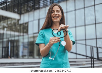 Smiling female nurse in teal scrubs holds a stethoscope shaped like a heart, symbolizing care and compassion in healthcare in an outdoor setting. - Powered by Shutterstock