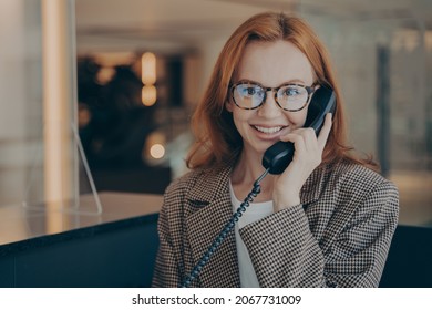 Smiling Female Office Worker With Red Hair In Plaid Jacket Wearing Glasses, Talking On Landline Phone With Her Co-worker About Project Completion Date, Office Environment In Blurred Background