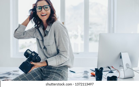 Smiling female photographer with a professional camera sitting on her desk. Woman with dslr camera in office looking away and smiling.