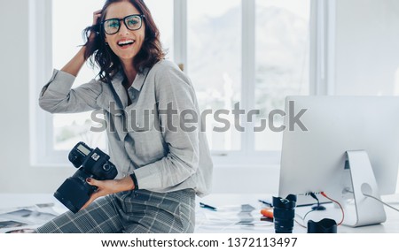 Smiling female photographer with a professional camera sitting on her desk. Woman with dslr camera in office looking away and smiling.