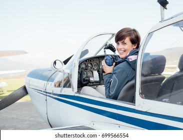 Smiling female pilot in the light aircraft cockpit, she is holding aviator headset and looking at camera