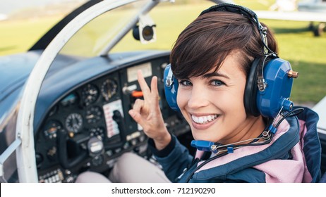 Smiling female pilot in the light aircraft cockpit, she is wearing aviator headset and making a V sign