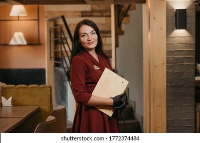 A Smiling Female Restaurant Manager In Black Disposable Medical Gloves Is Posing Holding A Wooden Menu In A Restaurant. A Kind Cafe Owner In A Ruby Dress.