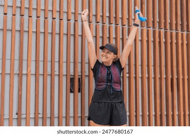 Smiling female runner in athletic gear celebrating with arms raised, standing against a wooden panel background. - Powered by Shutterstock