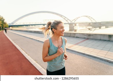 Smiling Female Runner With Blonde Hair And Pony Tail Dressed In Sportswear Running On The Kay Early In The Morning At Summertime.