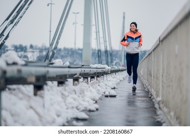 Smiling female runner jogging on a bridge during winter. Woman in sportswear running on the bridge on a snow. Copy space. Urban city jogger. Training in a urban area. - Powered by Shutterstock