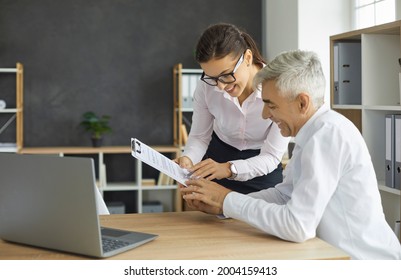 Smiling Female Secretary Gives Her Senior Manager Paper Documents Or A Contract To Sign. Happy Man Sitting At Office Desk And Reviewing Deal Or New Project. Cooperation Concept.