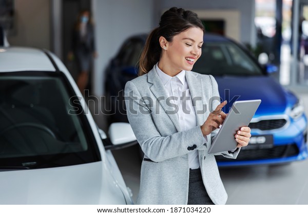 Smiling female seller in\
suit using tablet for looking which car is sold while standing in\
car salon.