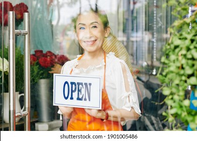 Smiling Female Senior Flower Shop Owner In Orange Apron Turning Open Sign On Glass Door