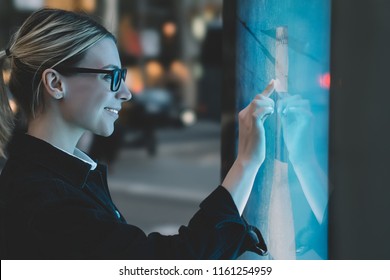 Smiling female standing at big display with advanced digital technology. Young woman touching with finger sensitive screen of interactive kiosk for find information while standing on street in evening - Shutterstock ID 1161254959