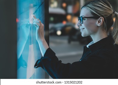 Smiling female standing at big display with advanced digital technology. Young woman touching with finger sensitive screen of interactive kiosk for find information while standing on street in evening