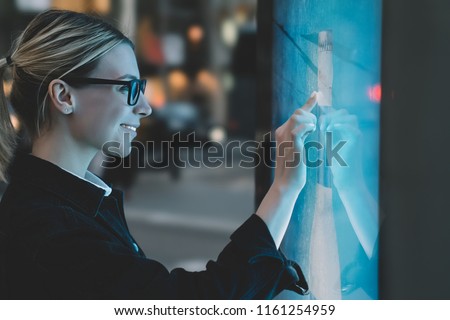 Smiling female standing at big display with advanced digital technology. Young woman touching with finger sensitive screen of interactive kiosk for find information while standing on street in evening