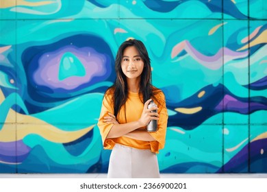 Smiling female street artist standing near the wall with her paintings holding spray paint can and looking to camera. Street art concept