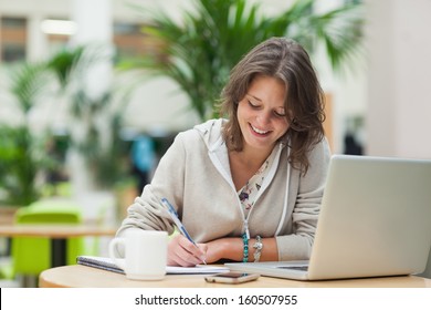 Smiling female student doing homework by laptop at cafeteria table - Powered by Shutterstock