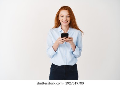 Smiling Female Student With Ginger Hair And Blue Eyes Using Mobile Phone, Looking Happy At Camera, Standing Over White Background.