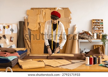 Smiling female tailor cutting pattern on large worktable in spacious fashion studio