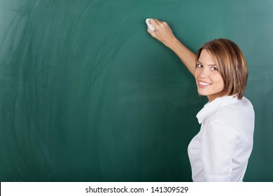 Smiling female teacher holding a chalk and writing on the blackboard - Powered by Shutterstock