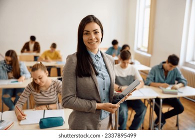 Smiling female teacher with touchpad during a class at high school looking at camera. Her student are learning in the background. - Powered by Shutterstock