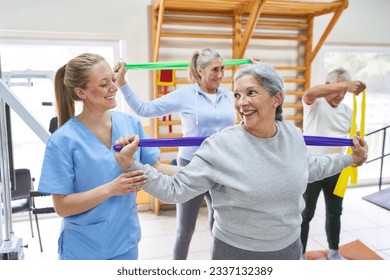 Smiling female therapist assisting elderly woman doing workout with resistance band at rehab center - Powered by Shutterstock