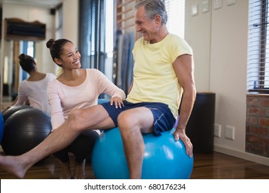 Smiling female therapist crouching by senior male patient sitting on exercise ball at hospital ward - Powered by Shutterstock