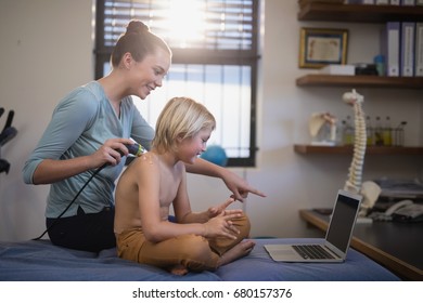 Smiling female therapist pointing at laptop while scanning shoulder of shirtless boy in hospital ward - Powered by Shutterstock