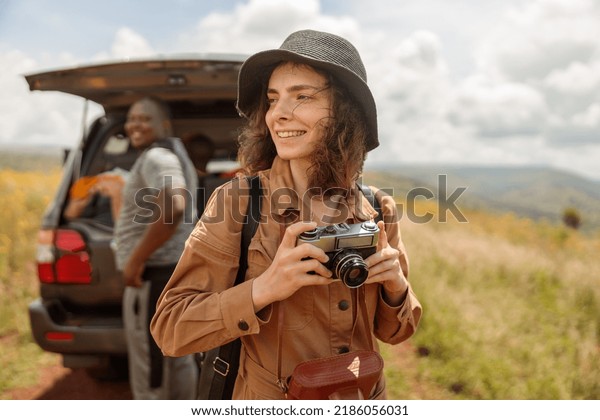 Smiling female traveler with\
photo camera enjoying the journey through the savannah next to\
safari car