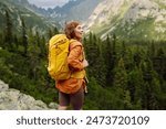 A smiling female traveler with a yellow hiking backpack enjoys the beautiful scenery of the majestic mountains. Travel, adventure. Concept of an active lifestyle.