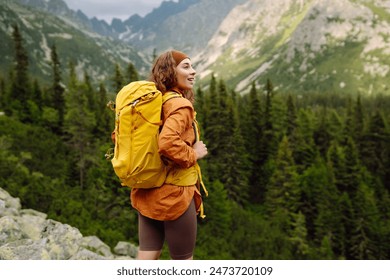 A smiling female traveler with a yellow hiking backpack enjoys the beautiful scenery of the majestic mountains. Travel, adventure. Concept of an active lifestyle.