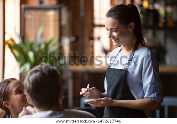 Smiling female waitress take order talk to\
clients cafe restaurant visitors couple, friendly professional\
woman server wear apron write dinner food menu choice, serving\
staff good customer\
service